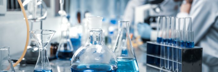 Laborsituation mit Erlenmeyerkolben im Vordergrund und Menschen mit weißen Kitteln im Hintergrund