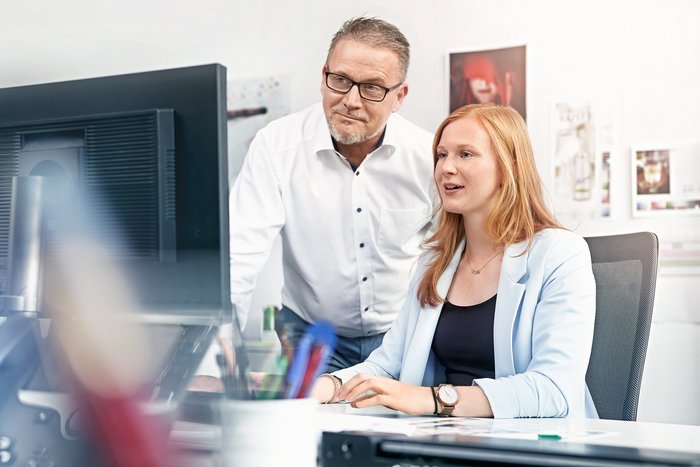 Eine junge Frau sitzt an Schreibtisch und sieht auf einen Monitor, ein Mann schaut ihr über die Schulter